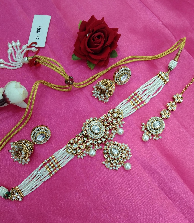 May include: An ornate gold-toned jewelry set featuring a choker necklace, earrings, and a headpiece, all adorned with white pearls and clear gemstones. The set is arranged on a pink surface, with a red rose and a tag visible.