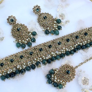 May include: A gold necklace set with green and white beads, a matching pair of earrings, and a tikka. The necklace is adorned with intricate floral designs and sparkling stones.