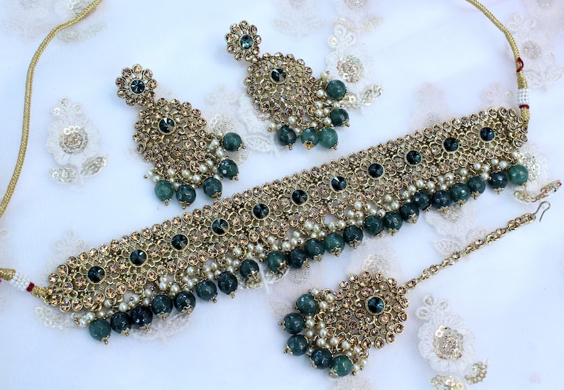 May include: A gold necklace set with green and white beads, a matching pair of earrings, and a tikka. The necklace is adorned with intricate floral designs and sparkling stones.