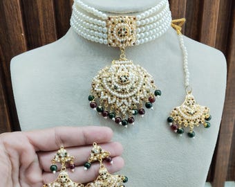 May include: A white pearl choker necklace with a gold pendant and matching earrings. The pendant features a circular design with maroon and green beads. The earrings are also circular with maroon and green beads. The text "Glam jewelrys" is visible at the bottom of the image.