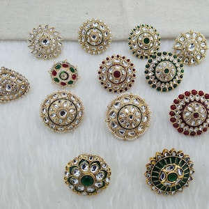 Puede incluir: Surtido de piezas de joyería redondas, decorativas, de color dorado con piedras de varios colores y detalles de perlas. Las piezas de joyería están dispuestas sobre un fondo de tela blanca.