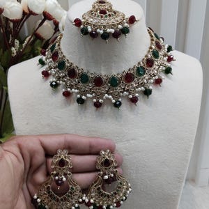 Puede incluir: Un conjunto de collar y pendientes de tono dorado con piedras rojas y verdes. El collar tiene un colgante con una piedra en forma de lágrima. El conjunto está etiquetado como "No. 1-Ruby Green" y "Glam jewelrys".