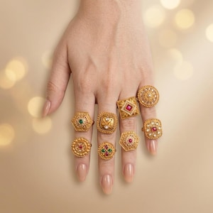 May include: A collection of eight gold-toned rings featuring intricate metalwork, small white pearls, and embedded red and green gemstones. The rings vary in shape, including circular, square, and hexagonal designs, all showcasing detailed filigree patterns.