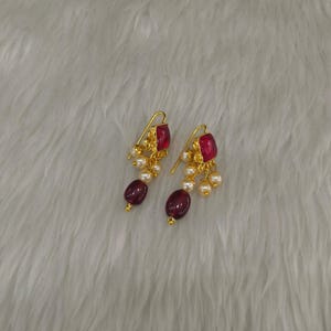 May include: A pair of gold-toned earrings featuring a square-cut deep red stone at the top, accented with small white pearls and a larger oval-shaped deep red bead dangling at the bottom, suspended from a curved gold-toned wire hook mechanism.