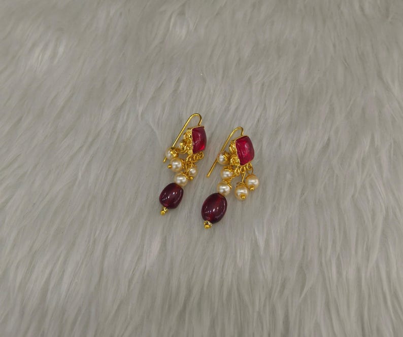 May include: A pair of gold-toned earrings featuring a square-cut deep red stone at the top, accented with small white pearls and a larger oval-shaped deep red bead dangling at the bottom, suspended from a curved gold-toned wire hook mechanism.