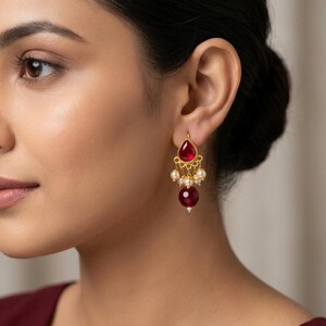May include: A gold-toned earring featuring a teardrop-shaped red stone at the top, a central section adorned with small white pearls, and a larger, round, deep red faceted bead dangling at the bottom, creating a layered, dangling design.