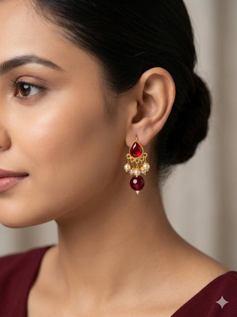 May include: A gold-toned earring featuring a teardrop-shaped red stone at the top, a central section adorned with small white pearls, and a larger, round, deep red faceted bead dangling at the bottom, creating a layered, dangling design.