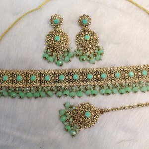 May include: A gold and green Indian bridal jewelry set with a choker necklace, earrings, and a maang tikka. The set features a delicate design with small, sparkling stones and beads.