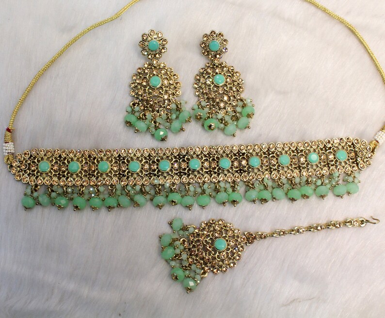 May include: A gold and green Indian bridal jewelry set with a choker necklace, earrings, and a maang tikka. The set features a delicate design with small, sparkling stones and beads.