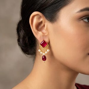 May include: A gold-toned earring featuring a square-cut deep pink gemstone at the top, suspended with a series of small white pearls and a single teardrop-shaped deep pink gemstone at the base, creating a dangling, multi-layered design.