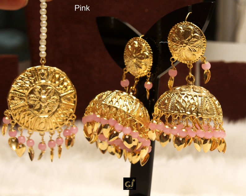 May include: A gold-toned jewelry set featuring a maang tikka with a white pearl strand and two jhumka earrings. Each piece displays intricate circular metal engravings, dangling pink beads, and small gold-colored leaf-shaped metal charms.