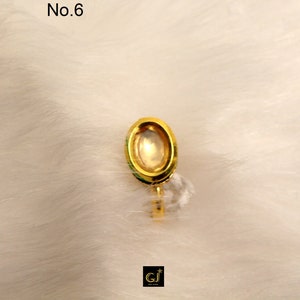 May include: A gold-colored oval-shaped ring with a clear stone in the center. The ring is on a white fuzzy surface. No. 6 is written in the top left corner of the image.