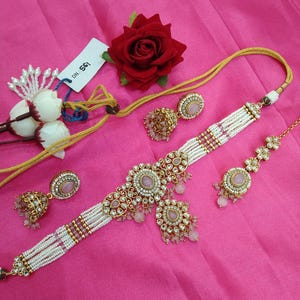 May include: An ornate jewelry set featuring a choker necklace, earrings, and a headpiece. The necklace and headpiece have pink gemstones and white beads. The earrings are gold-toned with pink stones. A red rose and white flowers are also in the image.
