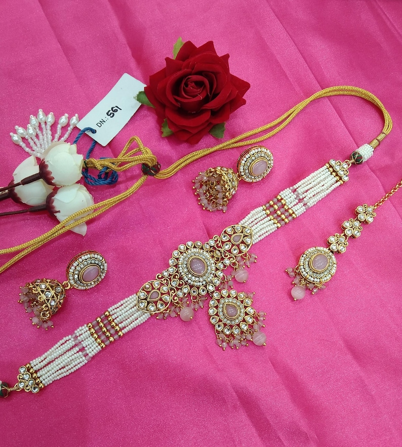 May include: An ornate jewelry set featuring a choker necklace, earrings, and a headpiece. The necklace and headpiece have pink gemstones and white beads. The earrings are gold-toned with pink stones. A red rose and white flowers are also in the image.