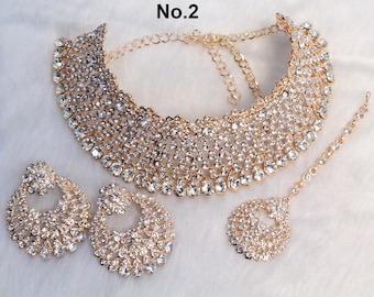 May include: A gold-tone bridal jewellery set with a large, multi-layered necklace, matching earrings, and a maang tikka. The set is adorned with clear rhinestones.