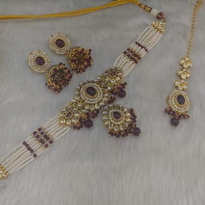 May include: An ornate jewelry set featuring a necklace, earrings, and a headpiece. The necklace and earrings have a gold-tone metal with white beads and dark red accents. The headpiece has a matching design. The set is displayed on a white, textured surface.