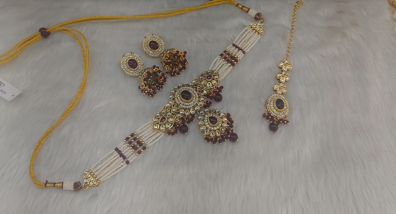 May include: An ornate jewelry set featuring a necklace, earrings, and a headpiece. The necklace and earrings have a gold-tone metal with white beads and dark red accents. The headpiece has a matching design. The set is displayed on a white, textured surface.