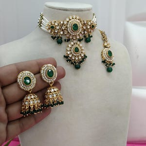 May include: A gold-toned jewelry set featuring a choker necklace, earrings, and a headpiece. The set is adorned with green gemstones and clear crystals. The choker has a white beaded base. The earrings are bell-shaped. The text "No. 2- Green" is visible.