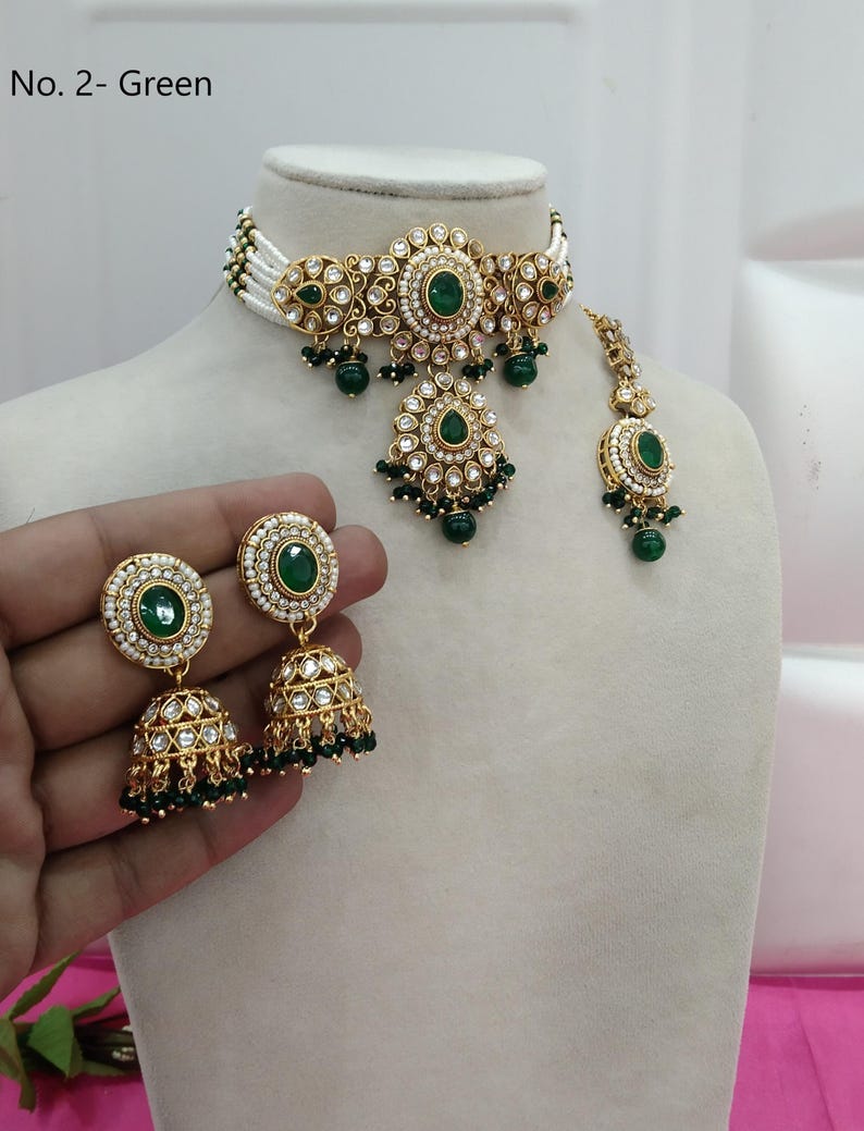 May include: A gold-toned jewelry set featuring a choker necklace, earrings, and a headpiece. The set is adorned with green gemstones and clear crystals. The choker has a white beaded base. The earrings are bell-shaped. The text "No. 2- Green" is visible.