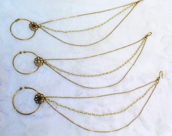 May include: Three gold nose rings with dangling chains. Each chain has three strands, two with small white beads and one with a gold chain. The nose rings have a decorative floral design with a red stone in the center.