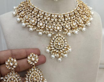 Peut inclure: Un ensemble de collier de mariée indien en or et blanc avec un grand pendentif, des boucles d'oreilles et une coiffe. Le collier est orné de perles et de pierres kundan.