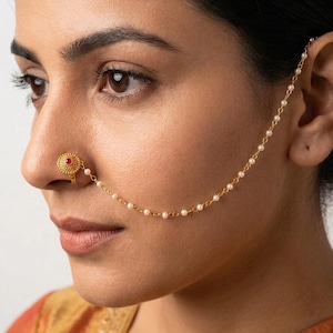 May include: A gold-colored nose ring with a small red gemstone in the center. The nose ring is connected to a chain of small white pearls that extends to the ear. The jewelry is worn on the left side of the face.