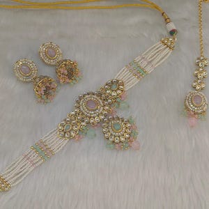 May include: An ornate jewelry set featuring a choker necklace, earrings, and a headpiece. The set includes gold-toned metal accents, pearls, and pink and green gemstones. The necklace and headpiece are connected by a delicate gold chain.
