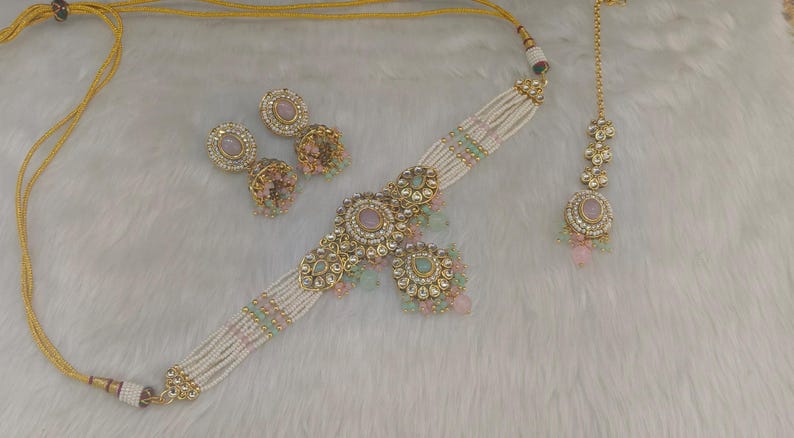 May include: An ornate jewelry set featuring a choker necklace, earrings, and a headpiece. The set includes gold-toned metal accents, pearls, and pink and green gemstones. The necklace and headpiece are connected by a delicate gold chain.