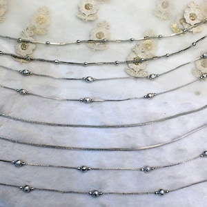 Könnte beinhalten: Ein Set aus acht silbernen Fußkettchen mit kleinen runden Perlen und dekorativen Anhängern. Die Fußkettchen sind in einer Reihe auf weißem Hintergrund angeordnet.