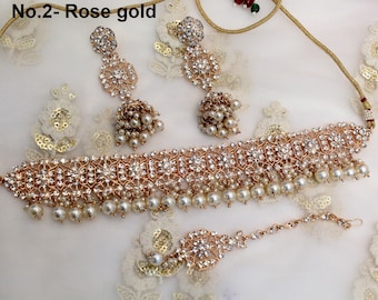 Op de afbeelding: Een roségouden choker-kettingset met een delicaat ontwerp met witte stenen en parels. De set bevat een bijpassend paar oorbellen en een maang tikka.