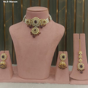May include: A maroon and gold-toned jewelry set displayed on a pink mannequin. The set includes a necklace, earrings, and a headpiece. The necklace features a central maroon stone surrounded by gold filigree and white beads. The earrings and headpiece mirror the necklace's design.