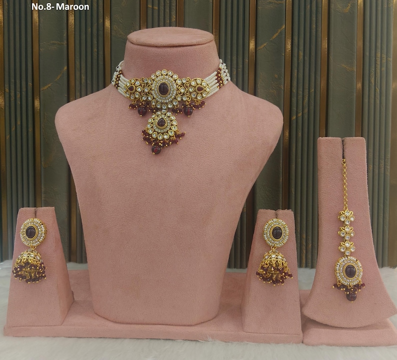 May include: A maroon and gold-toned jewelry set displayed on a pink mannequin. The set includes a necklace, earrings, and a headpiece. The necklace features a central maroon stone surrounded by gold filigree and white beads. The earrings and headpiece mirror the necklace's design.