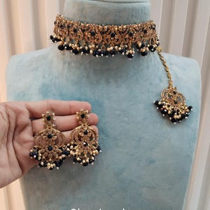 以下が含まれることがあります： 黒と白のビーズが付いたゴールドトーンのチョーカーネックレス、黒と白のビーズが付いたマッチングイヤリング、黒と白のビーズが付いたマッチングマングティッカ。セットには「No. 3-Blue」のラベルが付いています。