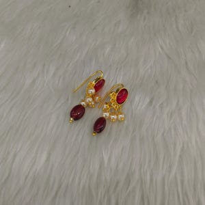 May include: A pair of gold-tone drop earrings featuring a single oval-shaped deep red stone at the base, accented by a cluster of small white pearls and a gold-tone hook wire for attachment. The design incorporates a metallic gold finish.