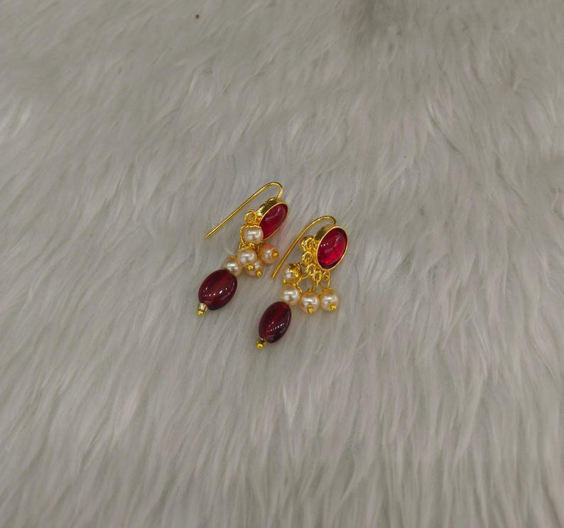 May include: A pair of gold-tone drop earrings featuring a single oval-shaped deep red stone at the base, accented by a cluster of small white pearls and a gold-tone hook wire for attachment. The design incorporates a metallic gold finish.
