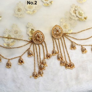 Puede incluir: Un par de pendientes de tono dorado con cadenas colgantes y detalles de perlas. Los pendientes presentan un diseño redondo y ornamentado con un motivo floral. Las cadenas están adornadas con pequeños amuletos en forma de campana.