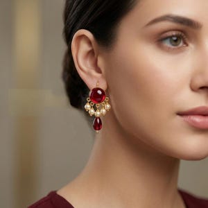 May include: A gold-toned earring featuring a circular red cabochon stone at the top, a row of small cream-colored spherical beads suspended below, and a single teardrop-shaped red stone dangling at the bottom center of the earring design.