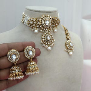 May include: A gold-toned jewelry set featuring a choker necklace, earrings, and a headpiece. The set includes white gemstones and small white pearls. The necklace and headpiece are adorned with intricate filigree designs. The earrings are bell-shaped.