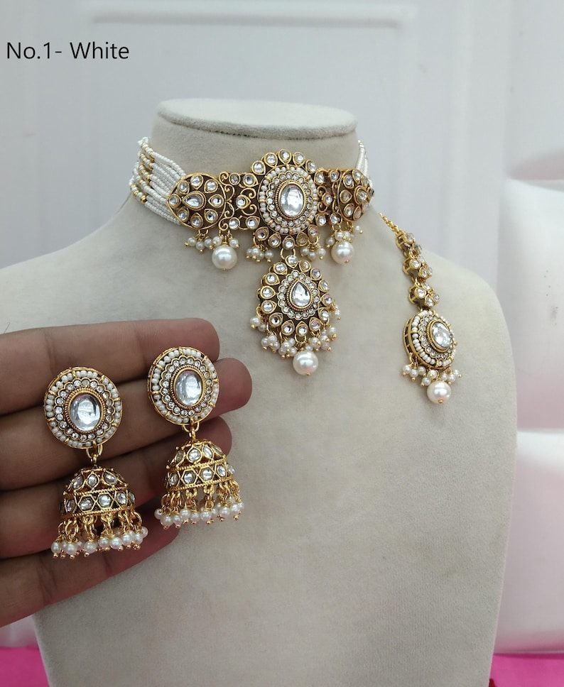 May include: A gold-toned jewelry set featuring a choker necklace, earrings, and a headpiece. The set includes white gemstones and small white pearls. The necklace and headpiece are adorned with intricate filigree designs. The earrings are bell-shaped.