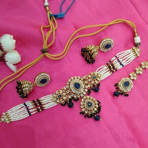 May include: An ornate jewelry set featuring a choker necklace, earrings, and a bracelet. The set includes gold-toned metal, white beads, and dark blue gemstones. The necklace and bracelet have a floral design.
