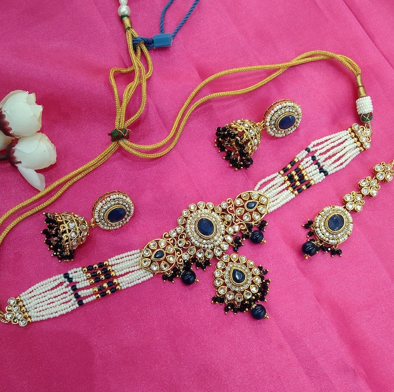 May include: An ornate jewelry set featuring a choker necklace, earrings, and a bracelet. The set includes gold-toned metal, white beads, and dark blue gemstones. The necklace and bracelet have a floral design.