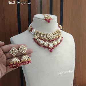May include: A gold-toned jewellery set featuring a choker necklace, a pair of jhumka earrings, and a maang tikka. The pieces are adorned with white stones, small white pearls, and magenta beads. The text No.2- Majenta is visible in the top left corner.