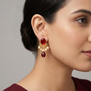 May include: A gold-toned earring featuring a round, deep red cabochon stone at the top, accented by a row of small, cream-colored spherical beads and a single, larger, teardrop-shaped red stone dangling from the center of the base.