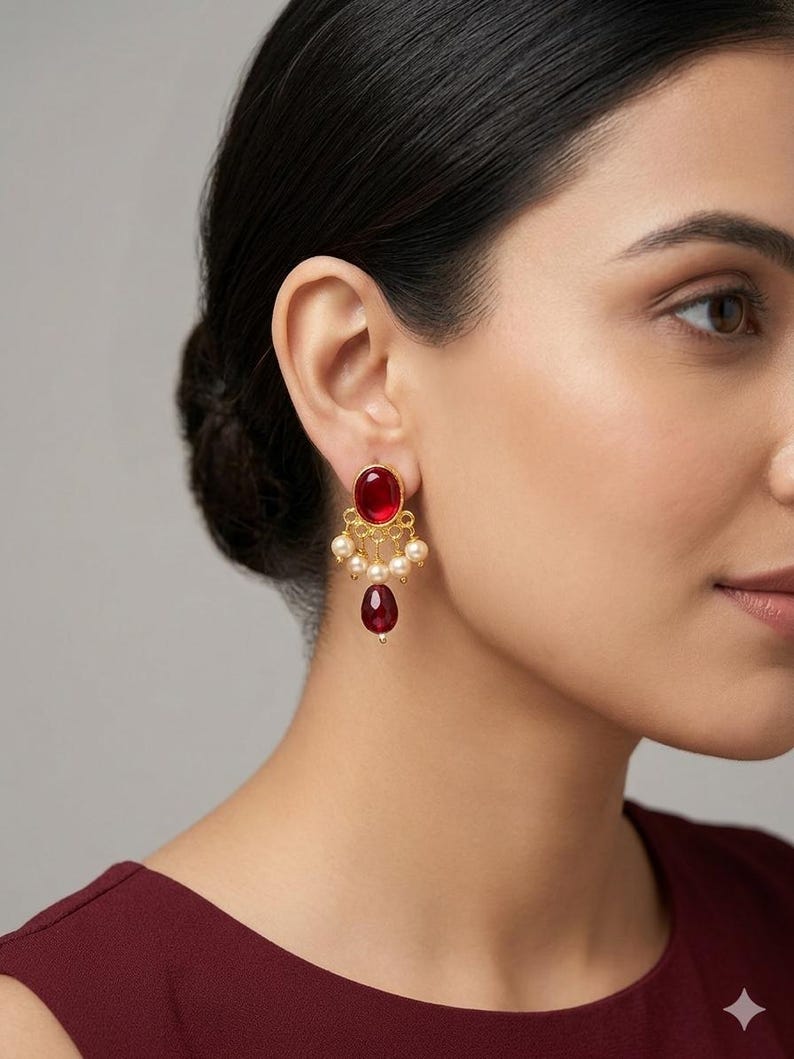 May include: A gold-toned earring featuring a round, deep red cabochon stone at the top, accented by a row of small, cream-colored spherical beads and a single, larger, teardrop-shaped red stone dangling from the center of the base.