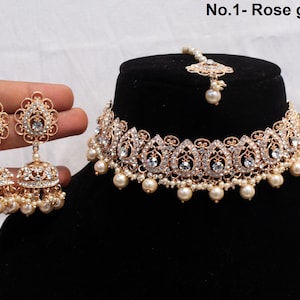 Puede incluir: Juego de joyas de novia en tono oro rosa con collar gargantilla, pendientes y tocado. El collar y los pendientes tienen un diseño floral con cristales transparentes y perlas de imitación. El tocado tiene una perla central.