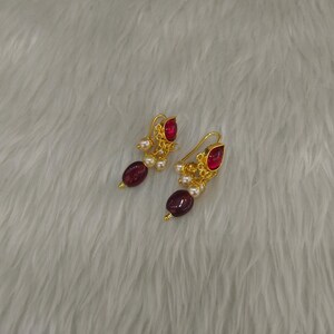 May include: A pair of gold-toned earrings featuring a teardrop-shaped red stone at the top, a cluster of small white pearls, and a larger oval-shaped dark red bead at the bottom, suspended from a curved gold-toned wire hook for pierced ears.