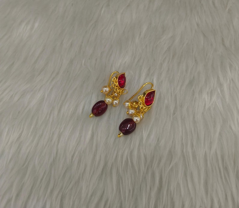 May include: A pair of gold-toned earrings featuring a teardrop-shaped red stone at the top, a cluster of small white pearls, and a larger oval-shaped dark red bead at the bottom, suspended from a curved gold-toned wire hook for pierced ears.