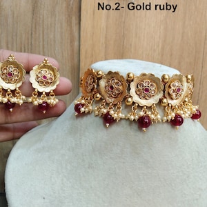 Op de afbeelding: Gouden choker ketting met robijnrode stenen en parel accenten. Passende oorbellen met robijnrode stenen en parel accenten. De ketting en oorbellen zijn gelabeld "No.2 - Gold ruby".