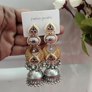 May include: A pair of gold and silver dangle earrings with a crescent moon design, pink and green stones, and a bell-shaped bottom. The earrings are on a white card with the text "fashion jewelry".