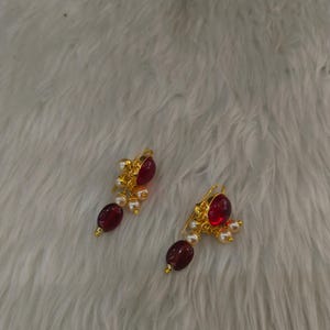 May include: A pair of gold-toned earrings featuring a large, oval-shaped deep red stone at the top, accented by smaller white pearls and a dangling dark red oval bead at the bottom, suspended from a gold-toned hook mechanism for ear attachment.